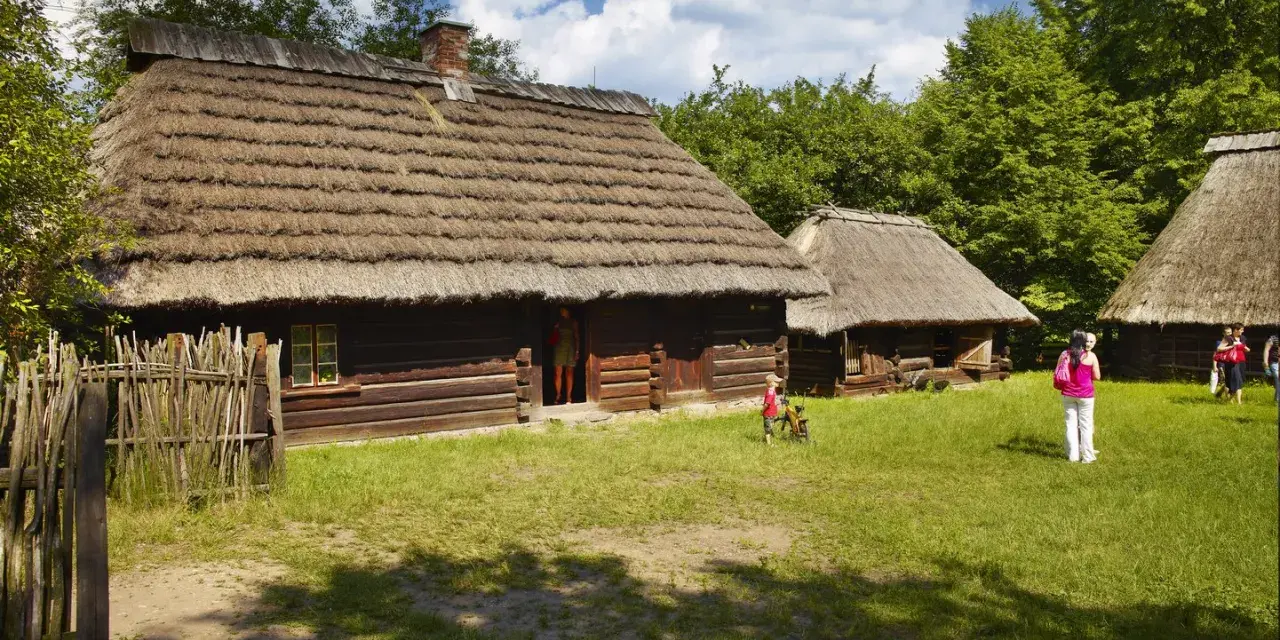 Skansen Chorzów bilety: drewniane chaty kryte strzechą, dzieci bawią się na trawie, zwiedzający podziwiają architekturę.