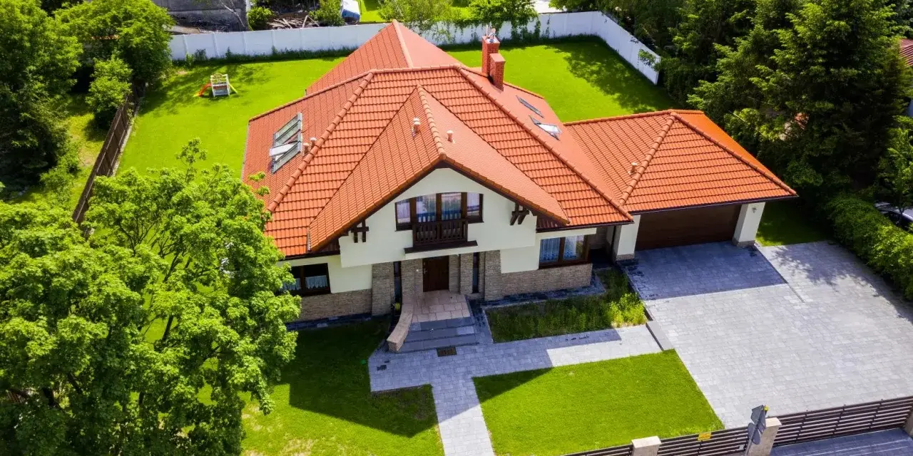 Piękny dom 200m2 z pomarańczowym dachem, otoczony zielonym ogrodem i drzewami. Podjazd z kostki brukowej prowadzi do garażu.