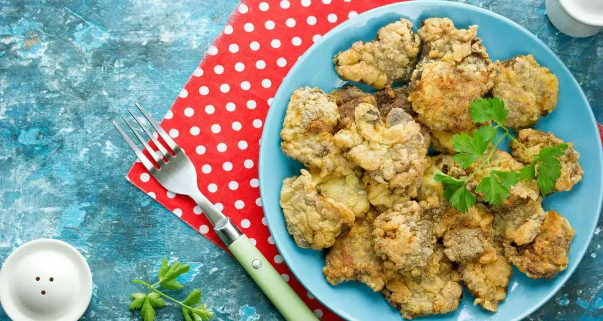 Piatto di fegatini di pollo impanati e fritti, guarniti con prezzemolo. Accanto, una forchetta e un sale e pepe.
