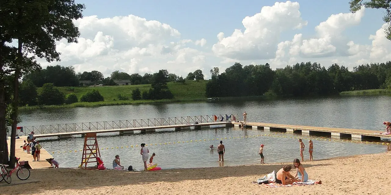 Słoneczne plaże na Mazurach tętnią życiem. Ludzie odpoczywają na piasku, kąpią się w jeziorze i spacerują po pomoście.