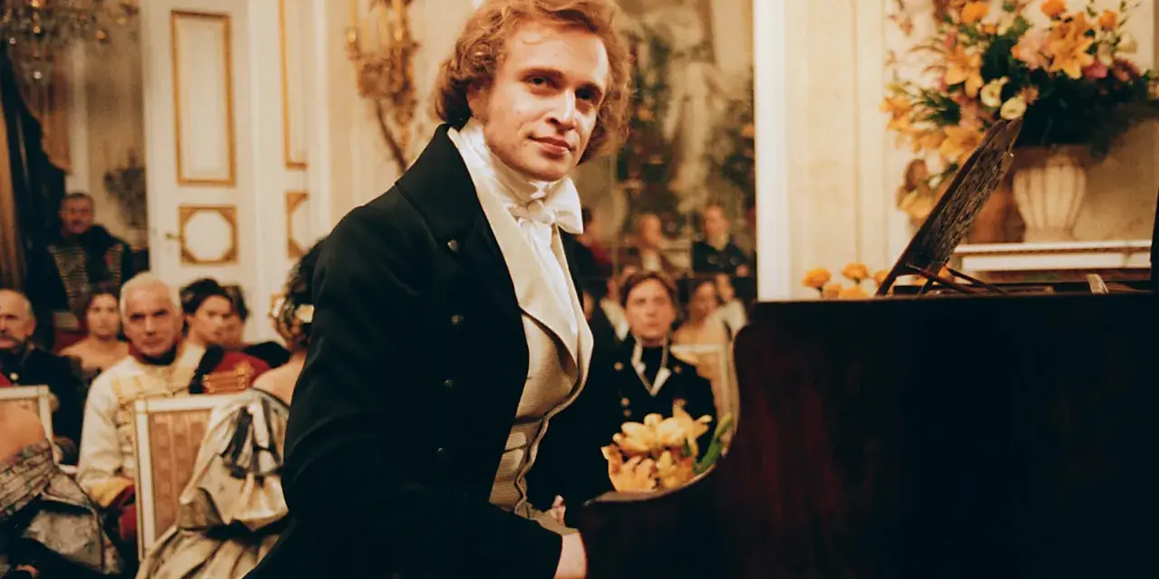 Piotr Adamczyk w historycznej scenie przy fortepianie, otoczony gośćmi w strojach z epoki. Jego role w filmach, serialach i programach zapadają w pamięć.