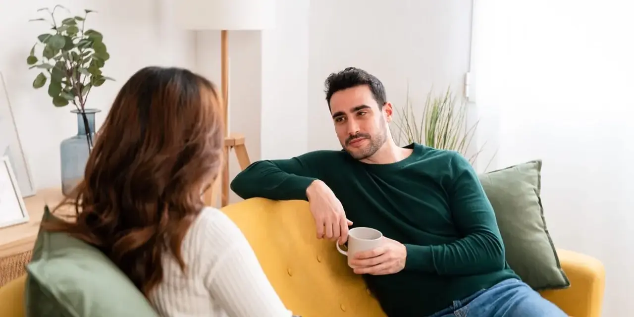 Coppia sul divano, lui con tazza in mano, lei di spalle. Una conversazione tesa, forse legata a **ADHD e tradimento**.