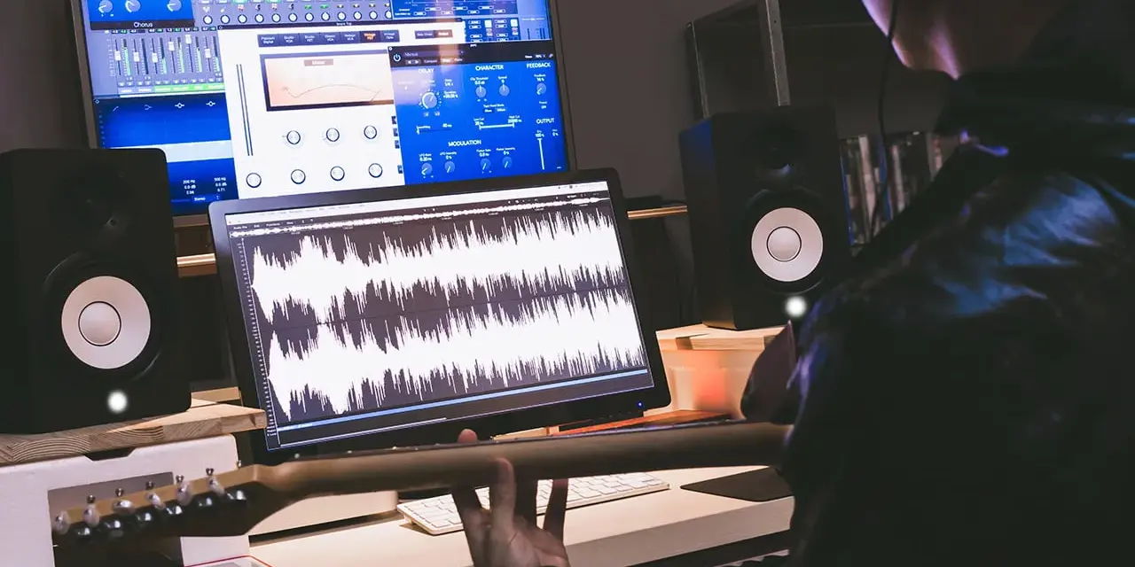 Tworzenie muzyki w domowym studiu. Na ekranach widać programy do edycji dźwięku, a w rękach gitarzysty gryf gitary.
