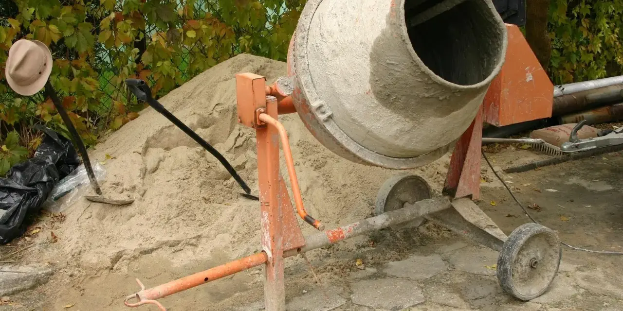 Betoniarka 200l czeka na piasek i cement. Ile cementu na betoniarkę 200l? Obok łopata i kapelusz.