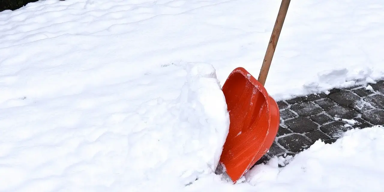 Pomarańczowa łopata do śniegu wbita w zaspę na ośnieżonym chodniku.