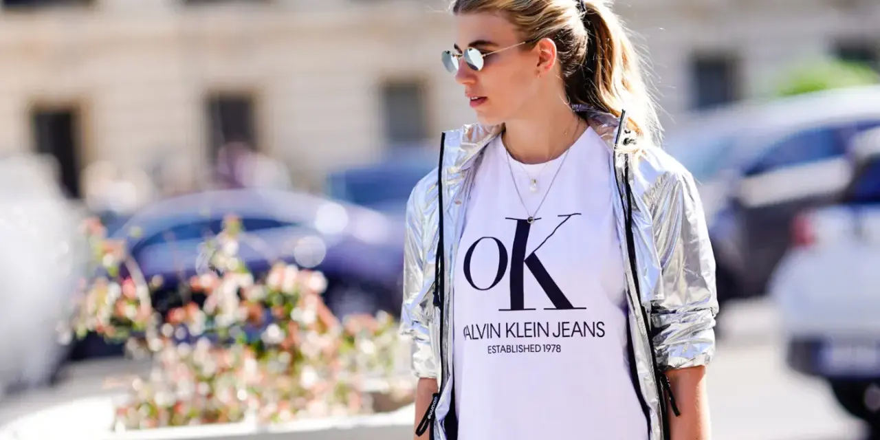 Kobieta w srebrnej kurtce CK damskiej i białej koszulce z logo Calvin Klein Jeans.