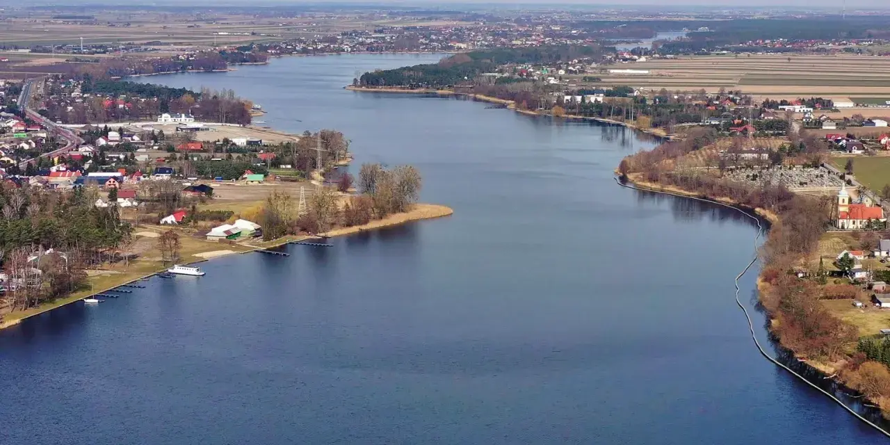 Rozległe jezioro Mikorzyńskie otoczone zabudowaniami i polami. W oddali widać kościół i cmentarz.