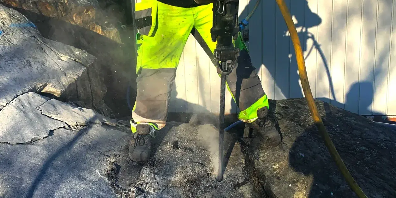 Arbetare med reflexkläder borrar i berg med en kraftig borrhammare. Dammet yr runt borren.