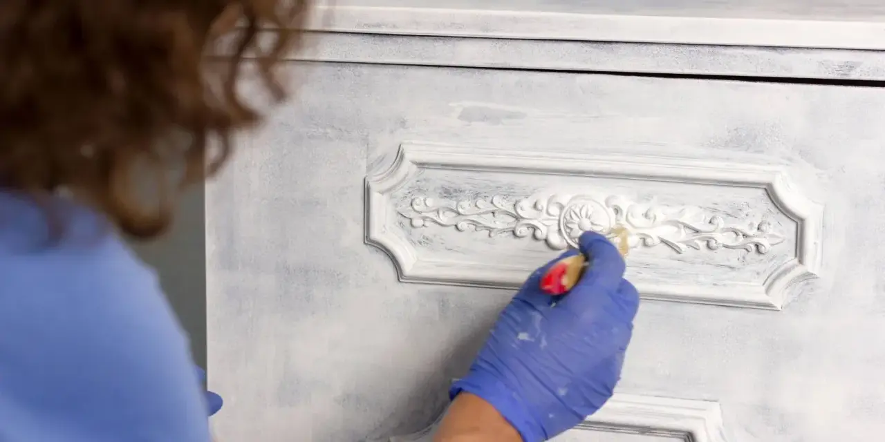 Mano enguantada aplica pintura a muebles pintados a la tiza en dos colores, dando un acabado desgastado y elegante.