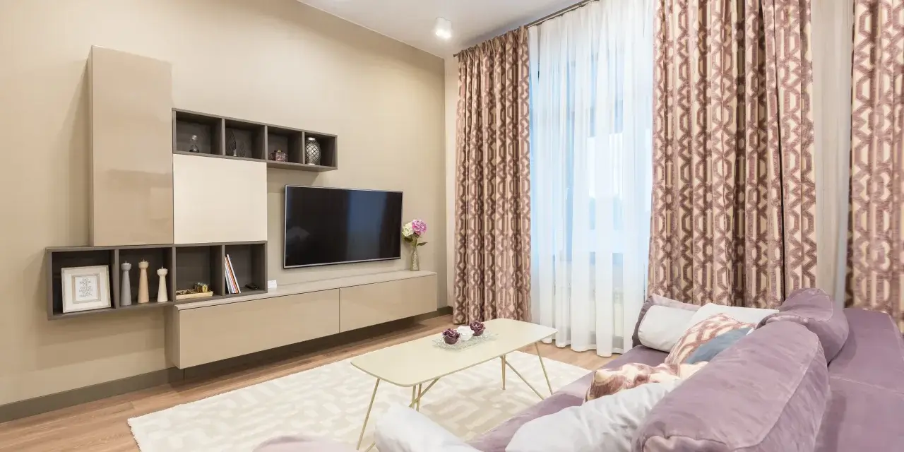 Przytulny salon z fioletową kanapą, beżowy kolor ścian tworzy ciepłą atmosferę. W tle telewizor i zasłony.