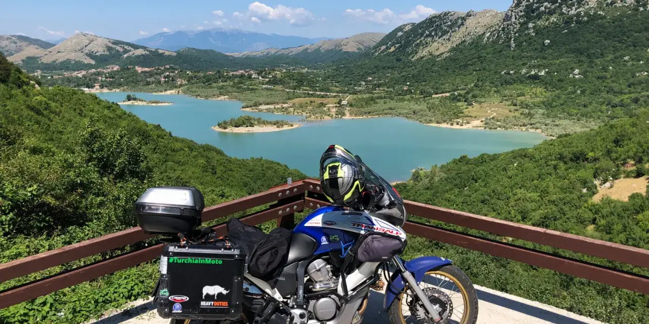 Moto pronta per avventurosi giri in moto nel Lazio, con un panorama mozzafiato sul lago turchese e le montagne verdi.