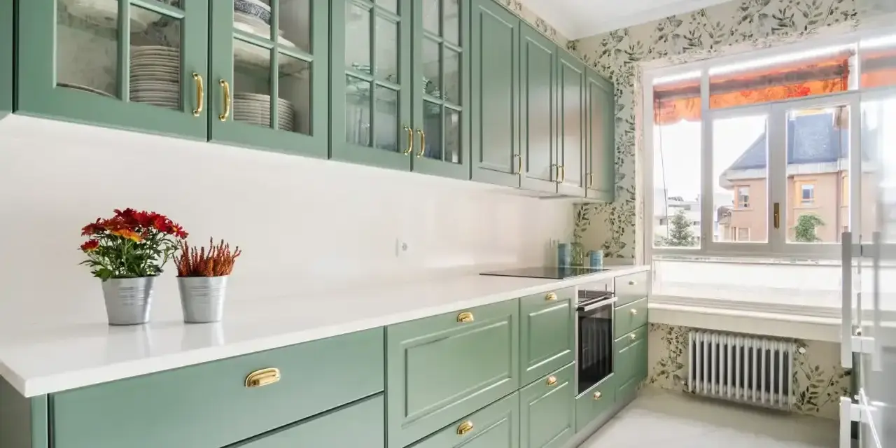 Encantadoras cocinas verdes vintage con armarios de cristal y detalles dorados. Flores rojas y naranjas adornan la encimera blanca.