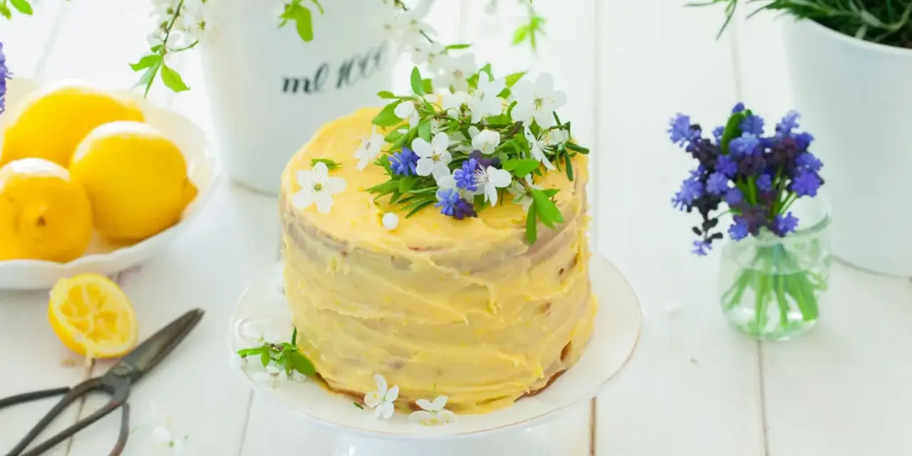 Słoneczny tort cytrynowy z kremem lemon curd, ozdobiony świeżymi kwiatami i ziołami. Obok leżą cytryny i stare nożyczki.