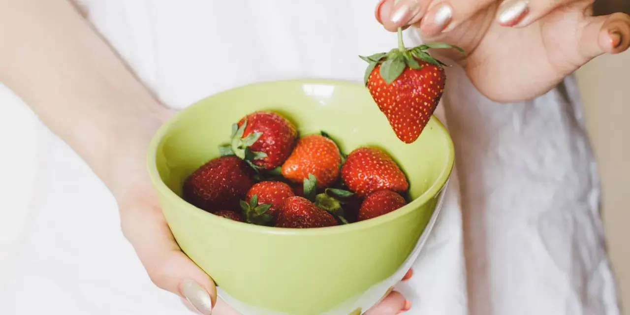 Hände halten eine hellgrüne Schüssel mit frischen Erdbeeren. Eine Erdbeere wird über der Schüssel gehalten, bereit zum Waschen.