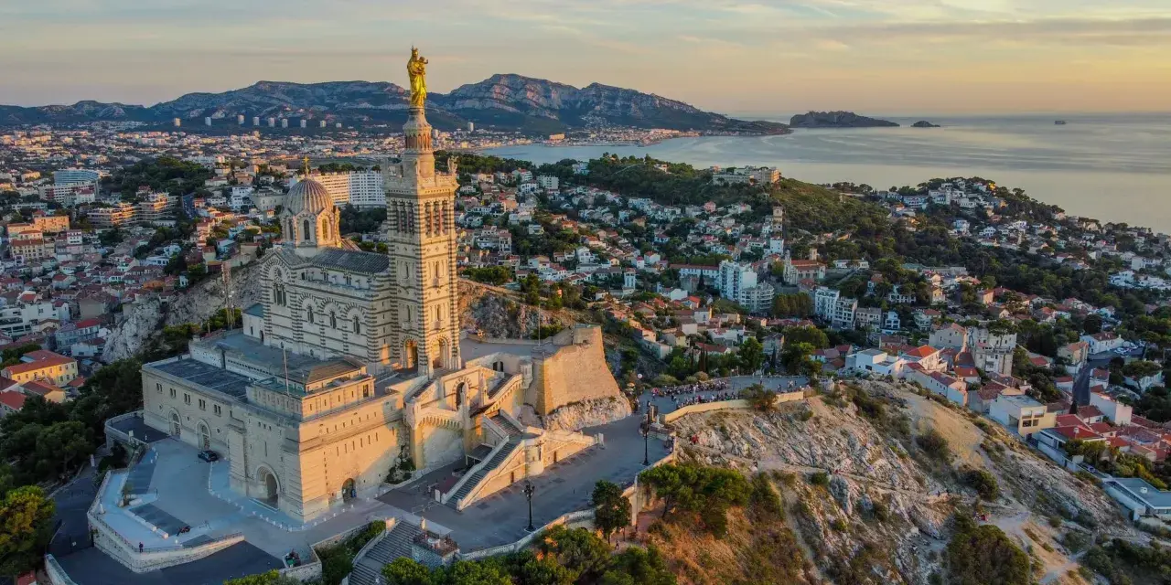 La basilique Notre-Dame de la Garde veille sur Marseille, baignée par la lumière dorée du soleil couchant. La mer et les collines environnantes s'étendent à perte de vue.
