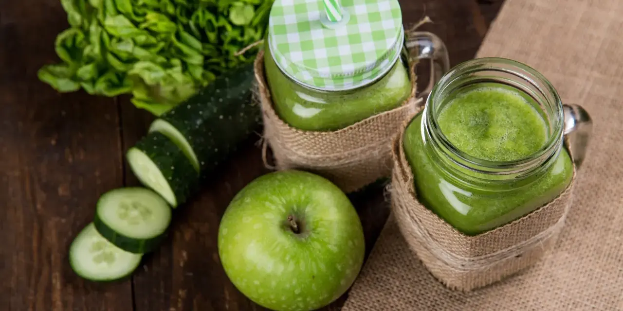 Dwa słoiki z zielonym smoothie, obok jabłko, plasterki ogórka i sałata. Orzeźwiający drink z ogórkiem na drewnianym tle.