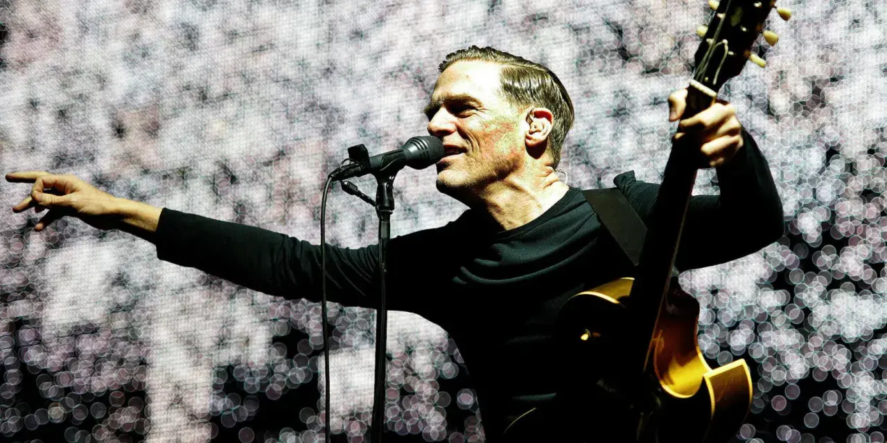 Bryan Adams w Polsce, śpiewa i gra na gitarze na tle świetlistej scenografii.