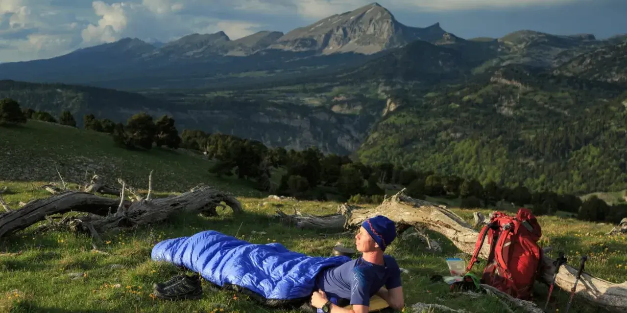 Mann liegt in blauer Biwakschachtel im Freien, umgeben von Berglandschaft. Rucksack und Wanderstöcke daneben.