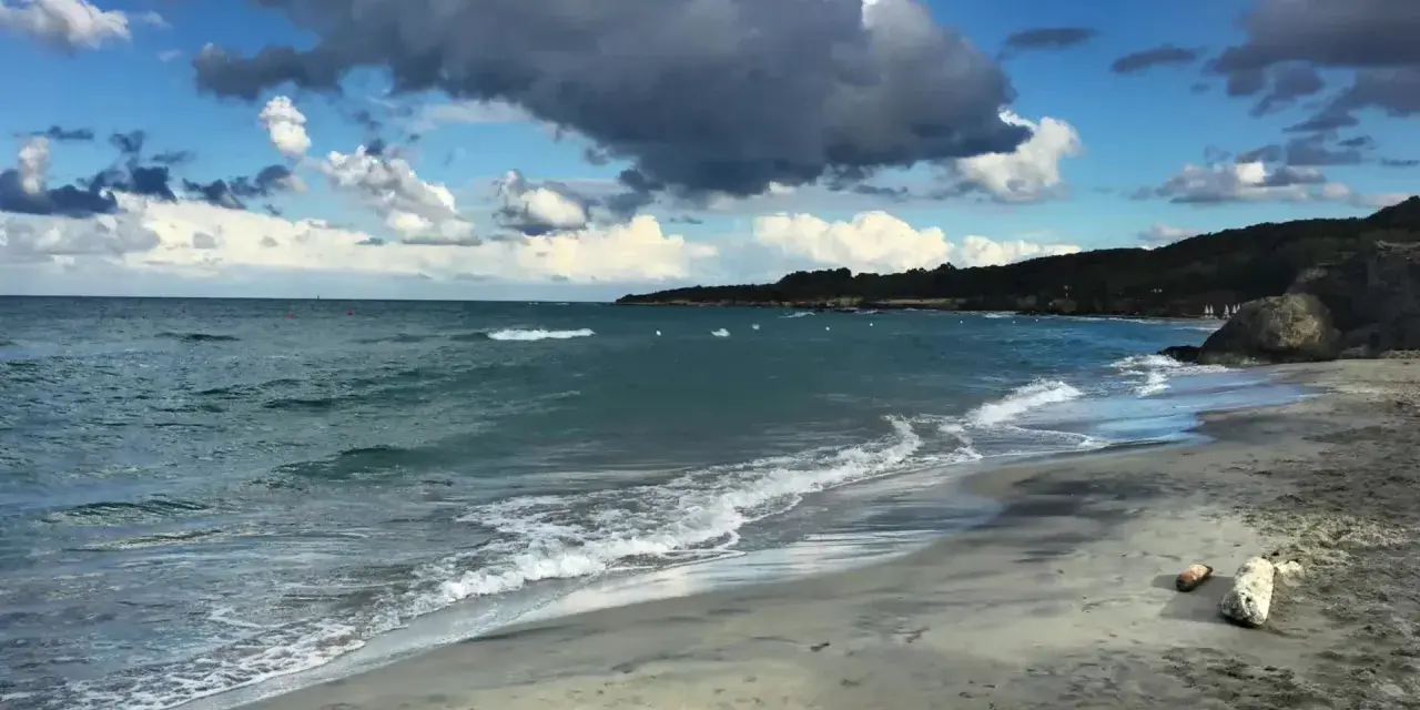 Onda che lambisce la spiaggia sabbiosa dell'Isola d'Elba, con il mare azzurro e nuvole scure nel cielo.