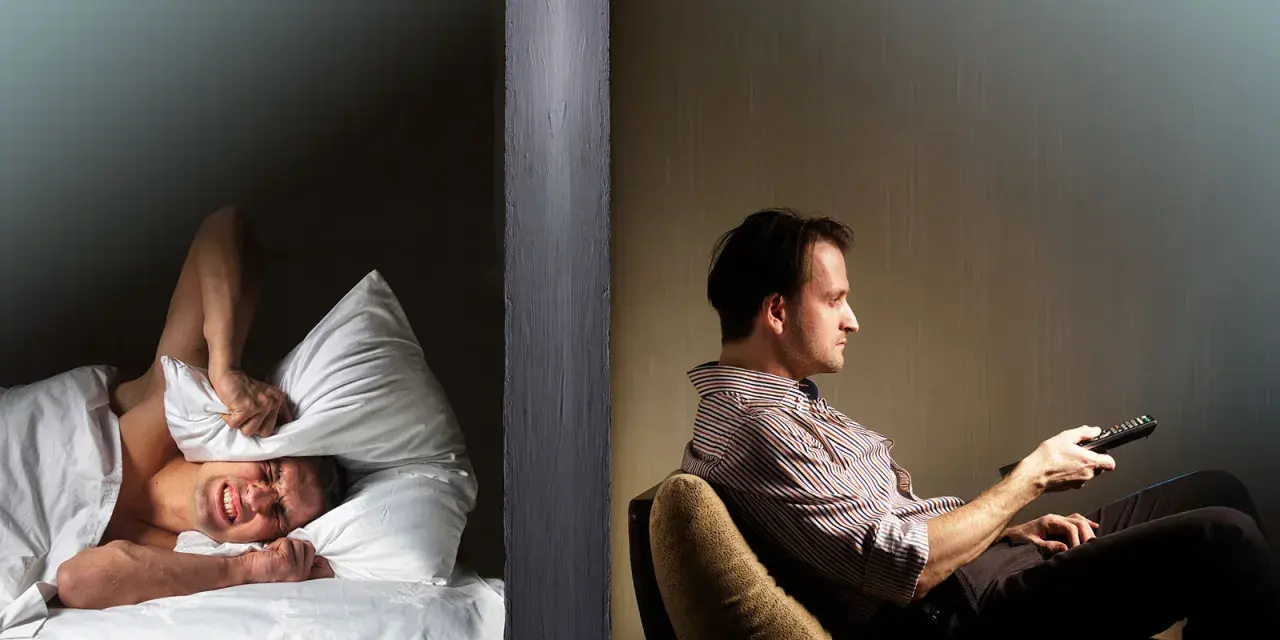 Hombre en cama tapa oídos con almohada, sufriendo por vecinos ruidosos. Al otro lado, otro hombre ve TV.