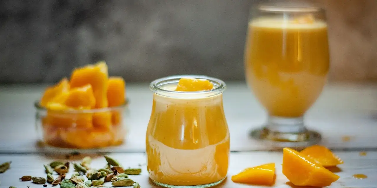 Deser z mango lassi kontra mus z marakui. Dwa pyszne, żółte desery w szklanych naczyniach, obok świeże kawałki mango i kardamon.