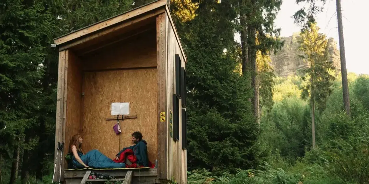 Zwei Personen entspannen sich in einer einfachen Holzhütte, bereit für wild camping. Umgeben von hohen Bäumen und üppigem Grün.