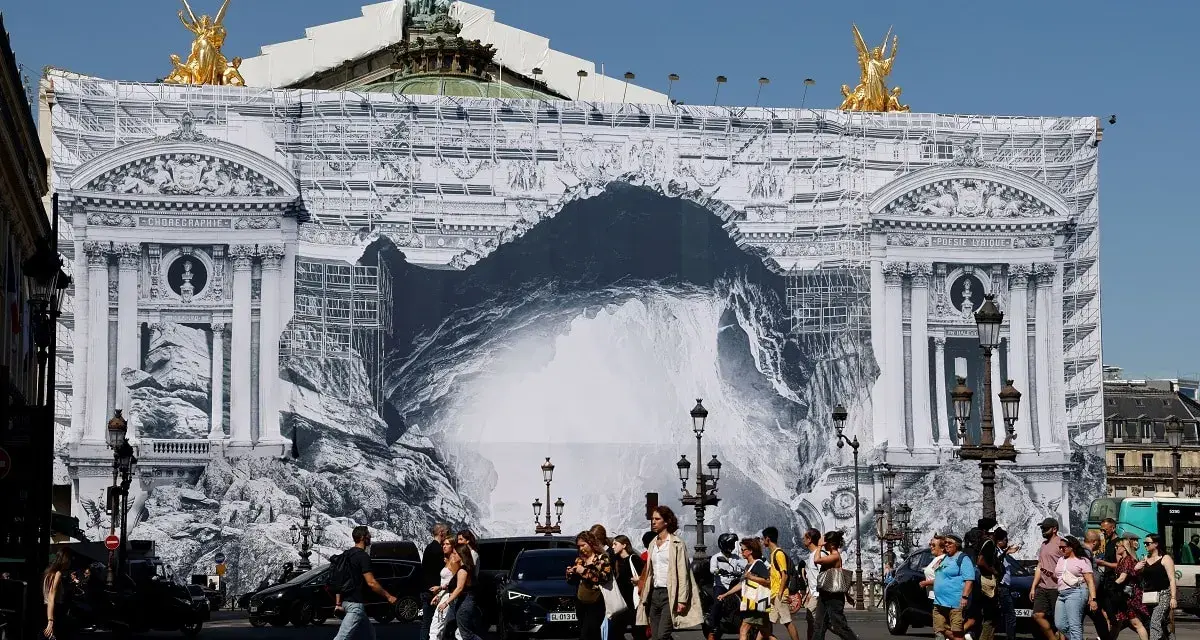 Des passants traversent devant l'Opéra Garnier, dont la façade est recouverte d'une bâche monumentale simulant des travaux et une arche rocheuse.