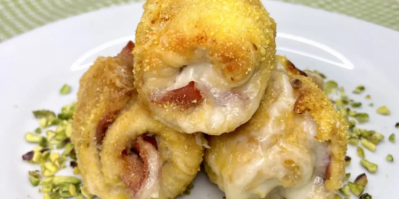 Tre involtini di pollo dorati e ripieni di formaggio filante e prosciutto, adagiati su un letto di pistacchi tritati.