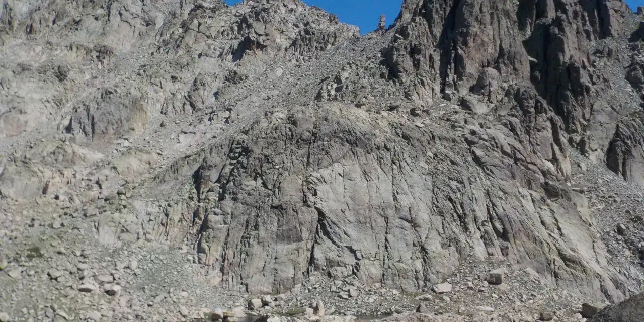 Vetta frastagliata di un monte rotondo, con rocce grigie e un lago alpino sullo sfondo.