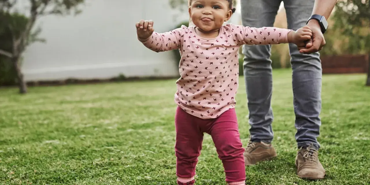Ein kleines Mädchen mit lustigen Zöpfen macht seine ersten Schritte im Gras, gehalten von einer erwachsenen Hand. Wann laufen Babys? Heute ist ein guter Tag dafür!