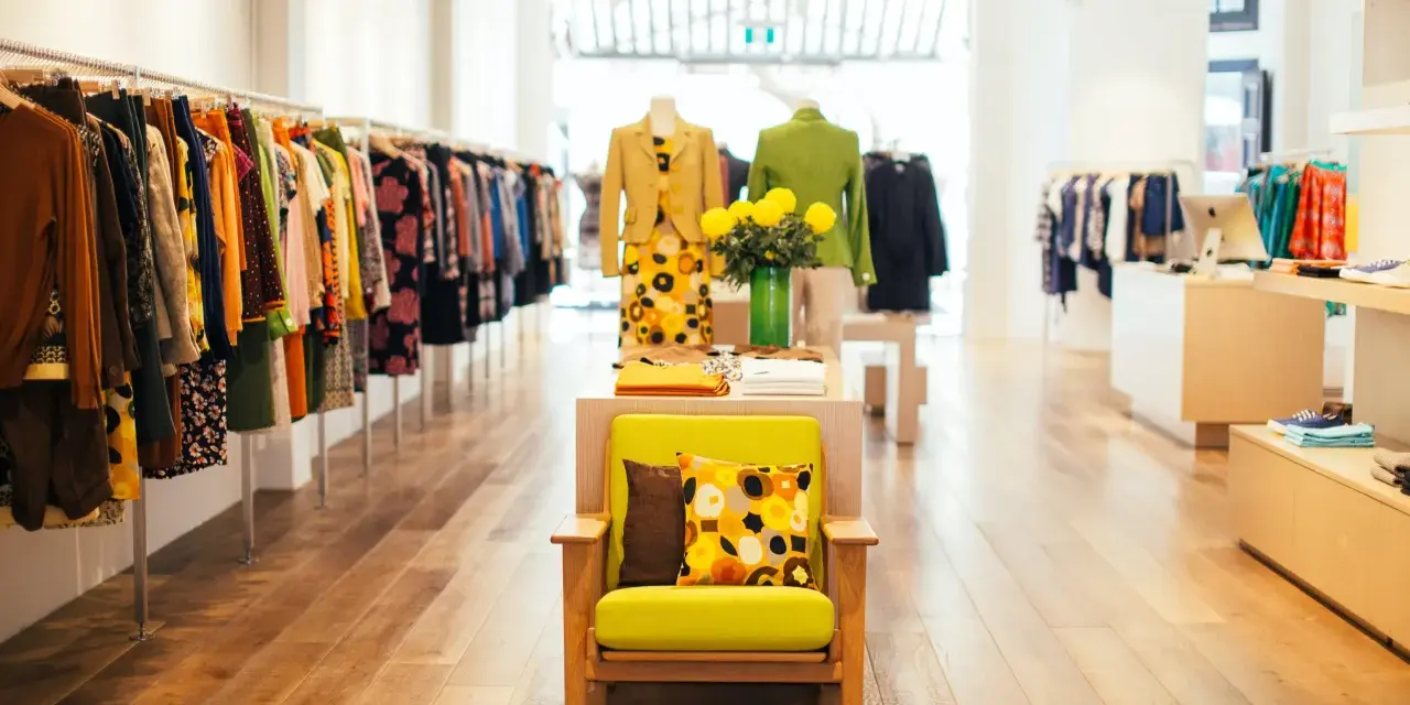 Interior de un showroom de ropa con percheros llenos de prendas coloridas y un sillón verde con cojines estampados.