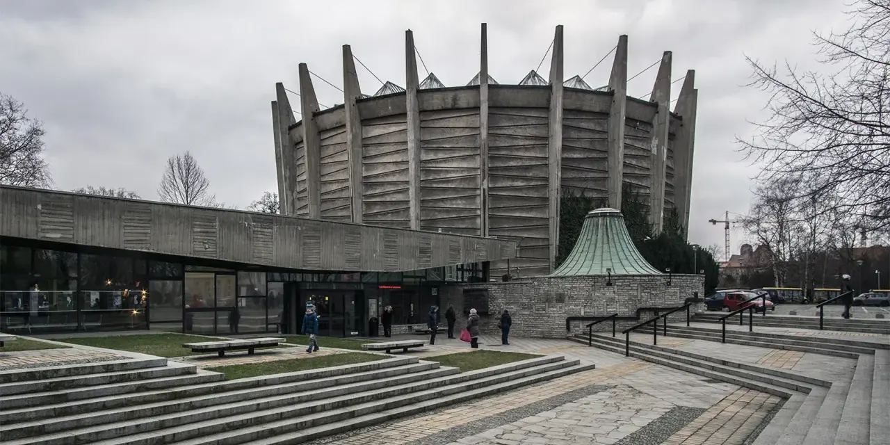 Monumentalny budynek w stylu brutalizm z betonowymi żebrami i szklaną fasadą. Schody prowadzą do wejścia, a wokół zielone trawniki.