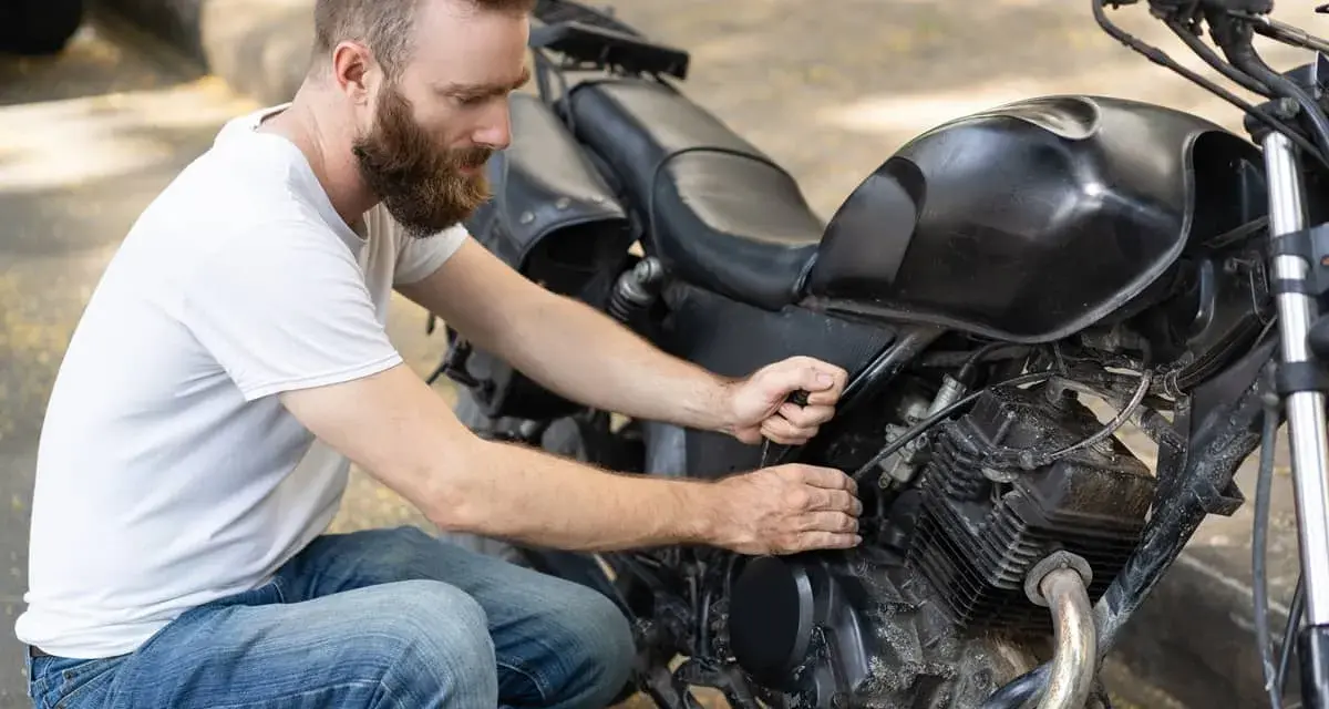 Uomo con barba lavora sul motore di una moto. Controlla i cavi, forse per il tempo di ricarica batteria moto 12v.