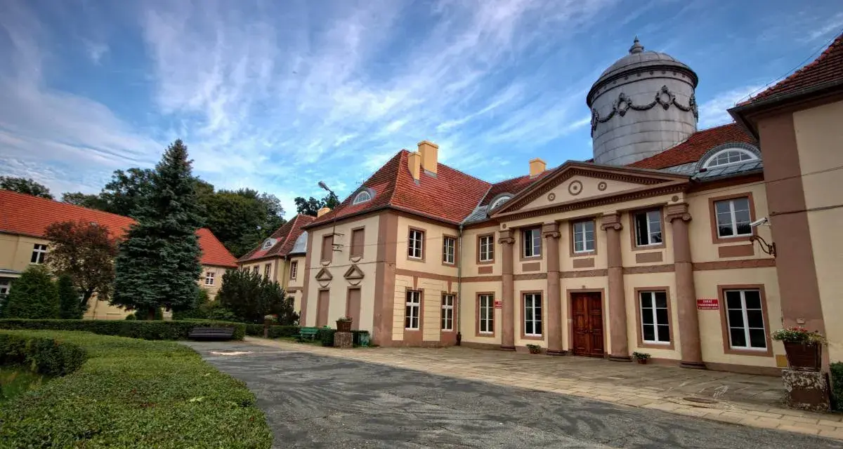 Pałac Maltzanów z charakterystyczną wieżą i czerwonym dachem, otoczony zielenią.