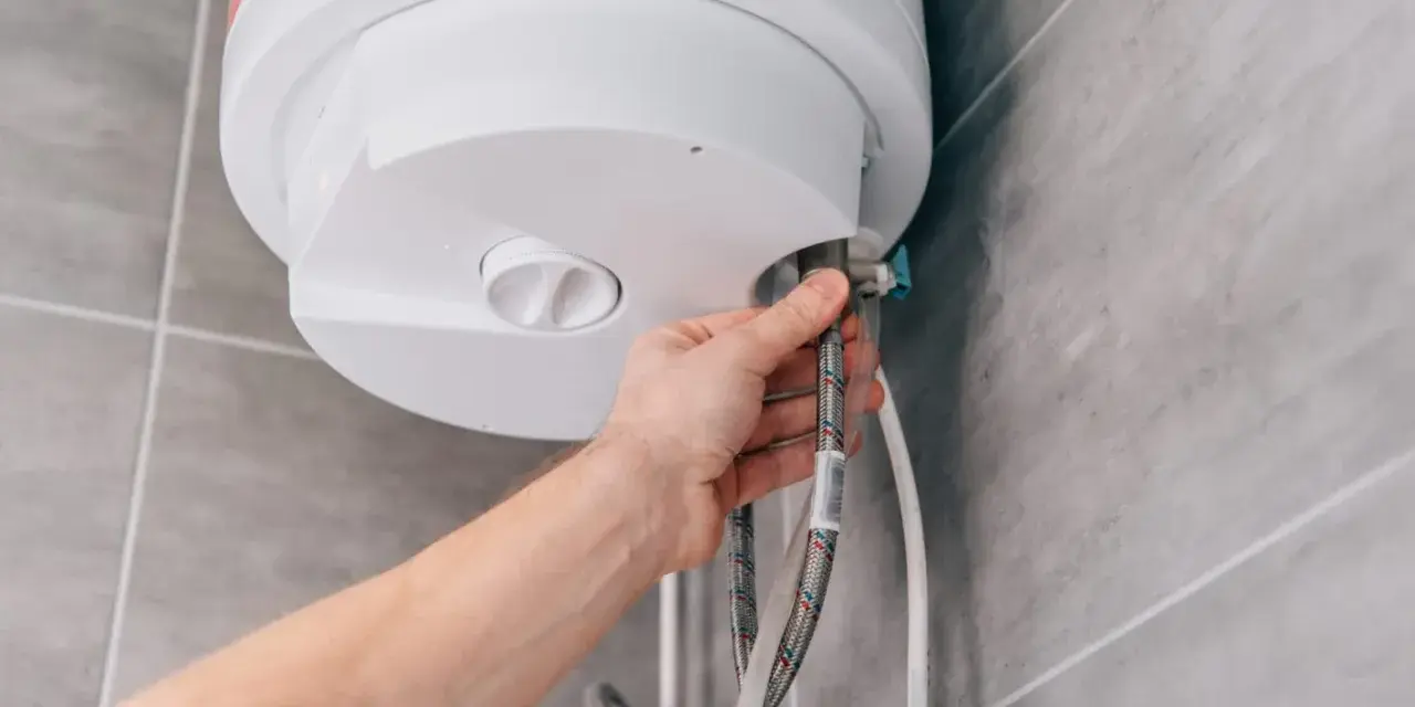 Mano che controlla la valvola scaldabagno perde. Tubi flessibili collegati a un boiler bianco su sfondo piastrellato.