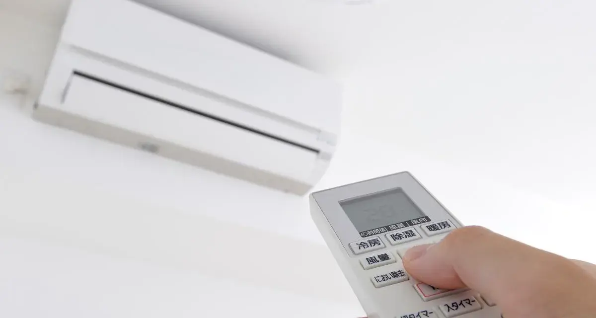 Mano ajustando el **timer on aire acondicionado** con el control remoto. El aire acondicionado blanco está montado en la pared.