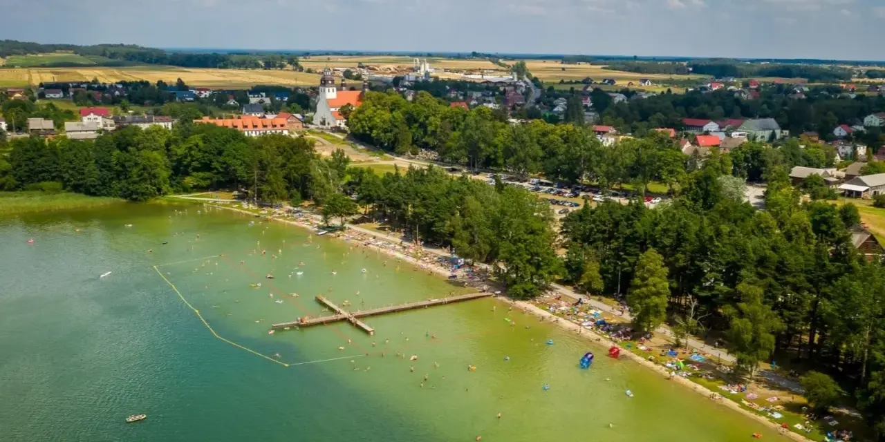 Letnie popołudnie nad jeziorem wielewskim. Tłumy plażowiczów, kąpielisko z pomostem i urokliwa miejscowość w tle.