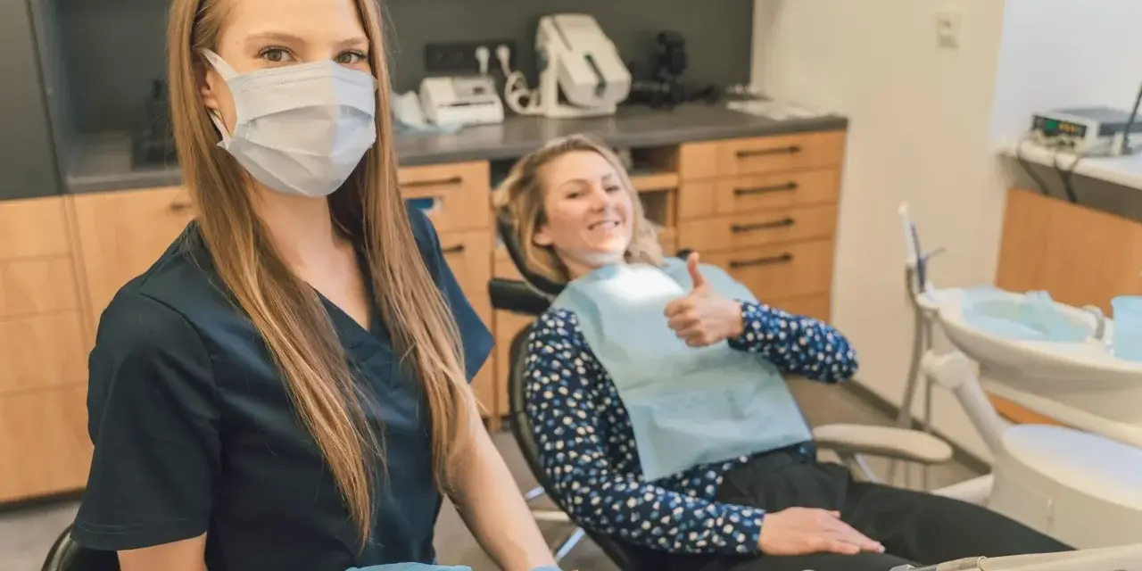 Uśmiechnięta pacjentka daje kciuk w górę podczas pierwszej wizyty u dentysty po latach. Dentystka w masce i rękawiczkach przygotowuje narzędzia.