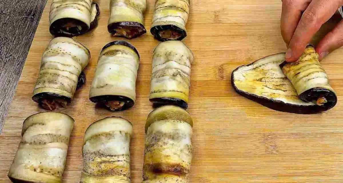Nove involtini di melanzane pronti, uno viene arrotolato su una fetta di melanzana grigliata.