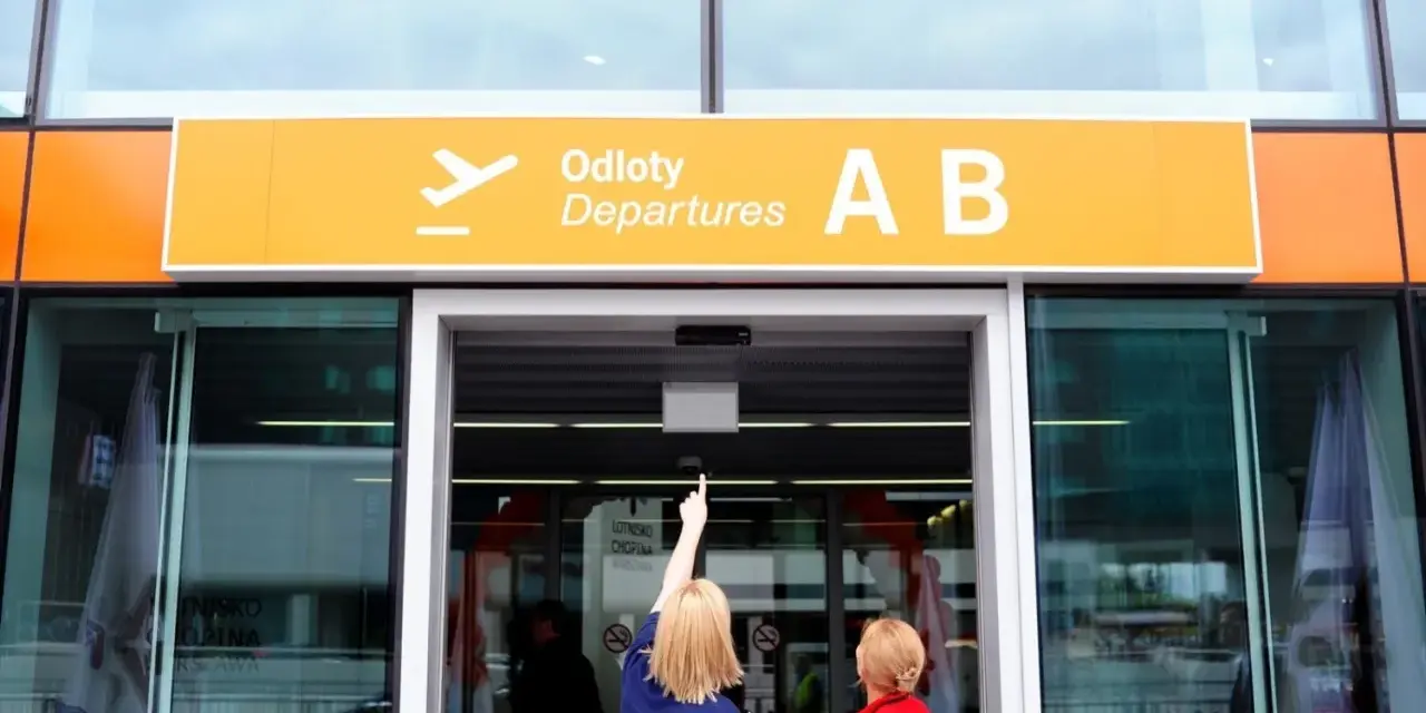 Dwie kobiety patrzą na tablicę "Odlocoty A B" na Lotnisku Chopina. Zastanawiają się, ile kosztuje parking na lotnisku Chopina.