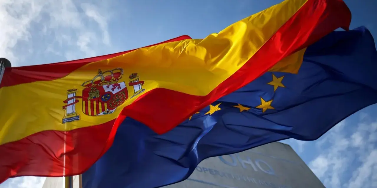 Flaga Hiszpanii, czerwono-żółta, powiewa obok flagi Unii Europejskiej na tle błękitnego nieba.