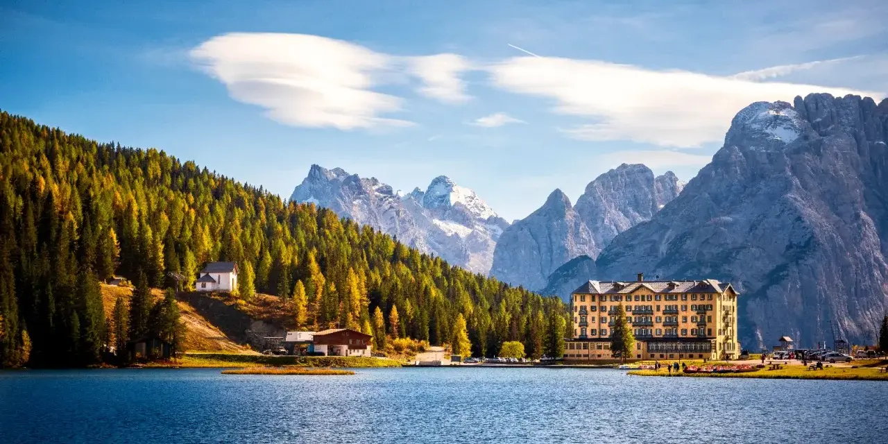 Laghi del Veneto in moto: un hotel giallo e una chiesetta bianca su un pendio boscoso, con montagne innevate sullo sfondo.
