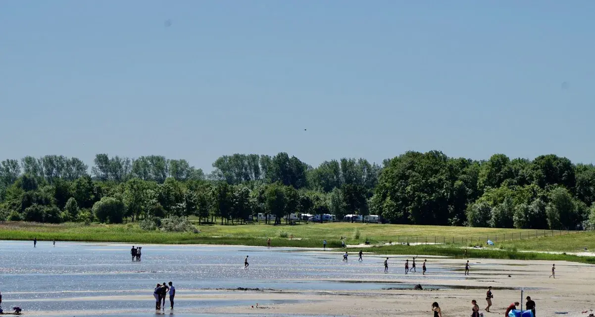 Menschen genießen einen sonnigen Tag am Strandbad Eldena. Einige baden im flachen Wasser, andere spazieren am Ufer entlang.