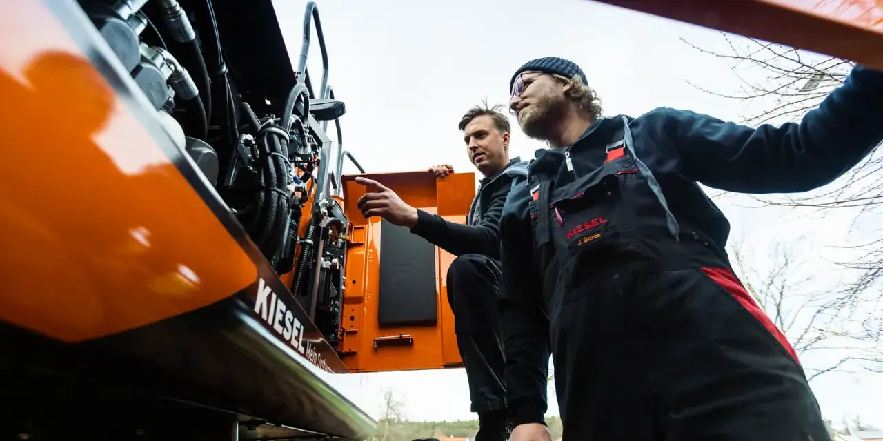 Dwóch mechaników w kombinezonach przeprowadza konserwację maszyn budowlanych. Jeden z nich wskazuje na silnik.