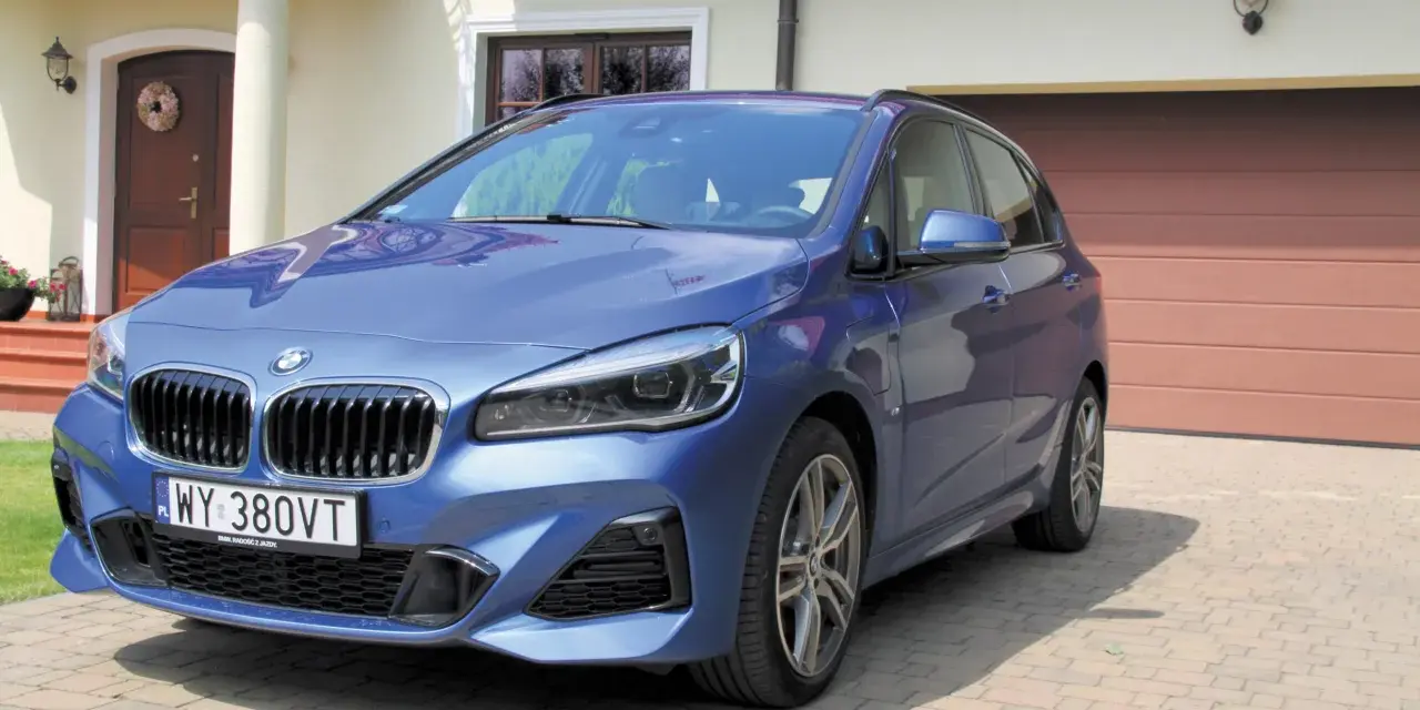 Niebieskie BMW 218i zaparkowane przed domem z brązową bramą garażową.