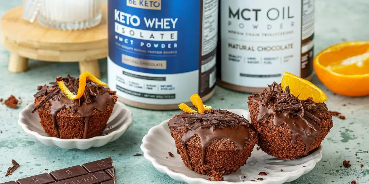 Pyszne keto muffiny z mąki migdałowej, polane czekoladą i ozdobione skórką pomarańczową. Obok stoją puszki z odżywką białkową i olejem MCT.