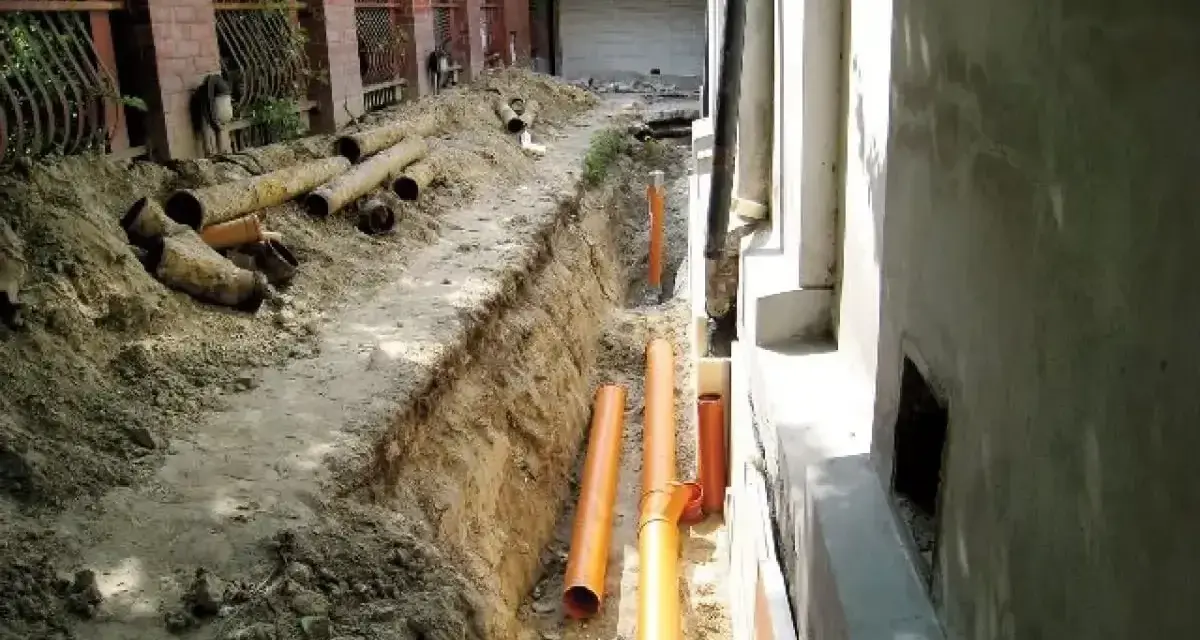Wykop pod drenaż wokół starego domu, widoczne nowe pomarańczowe rury i stare, zniszczone rury.