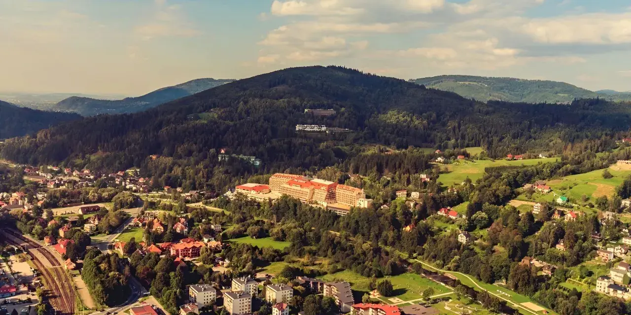 Widok z kamery na hotel Gołębiewski w Wiśle, otoczony zielonymi wzgórzami i zabudowaniami.