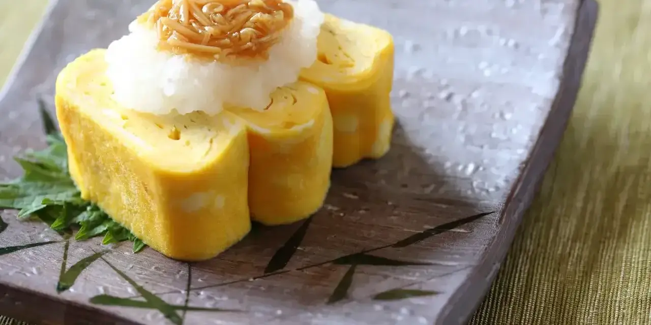 Delikatny omlet tamago, zwinięty w rolki, podany z tartą rzodkwią i grzybami enoki na talerzu z bambusowym wzorem.