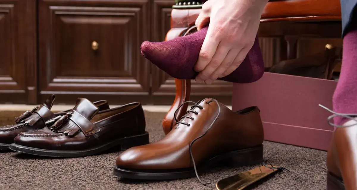 Hombre se pone calcetín morado para ablandar zapatos con crema hidratante. Zapatos de vestir y mocasines en el suelo.